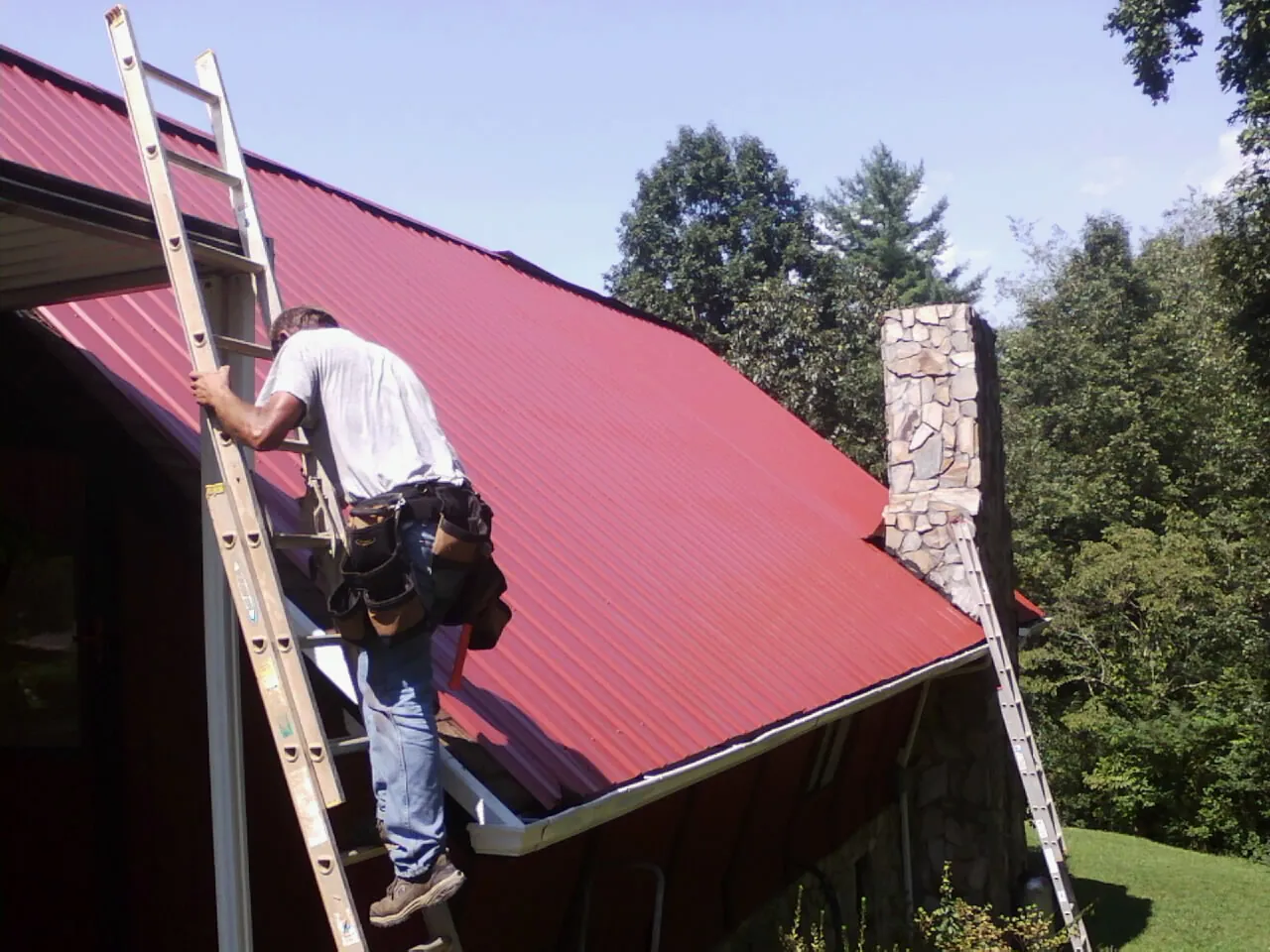 Roofer on a ladder beside a red metal roof and stone chimney — Soffit & Fascia Repair by Roofing Brothers in Merchantville